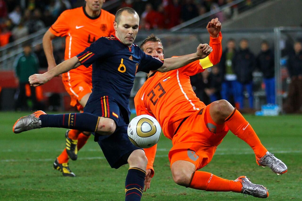 Two soccer players in a mid-match tackle during a game, one wearing a dark blue uniform, the other in bright orange, compete fiercely for the ball.