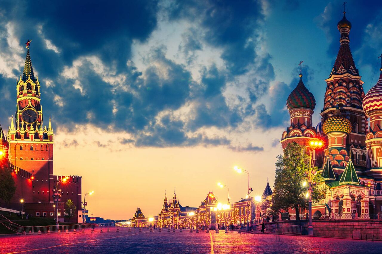 Sunset view of the historic Saint Basil's Cathedral and Spasskaya Tower in Moscow, Russia, lit up, highlighting their colorful architecture against a cloudy sky.