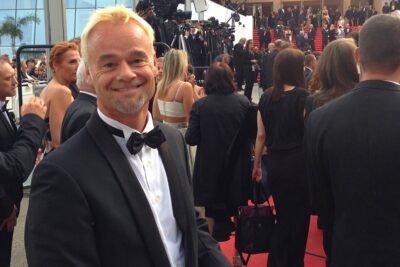 A man in a tuxedo smiles at the camera during a red carpet event, amid other formally dressed guests and photographers in a glamorous setting.