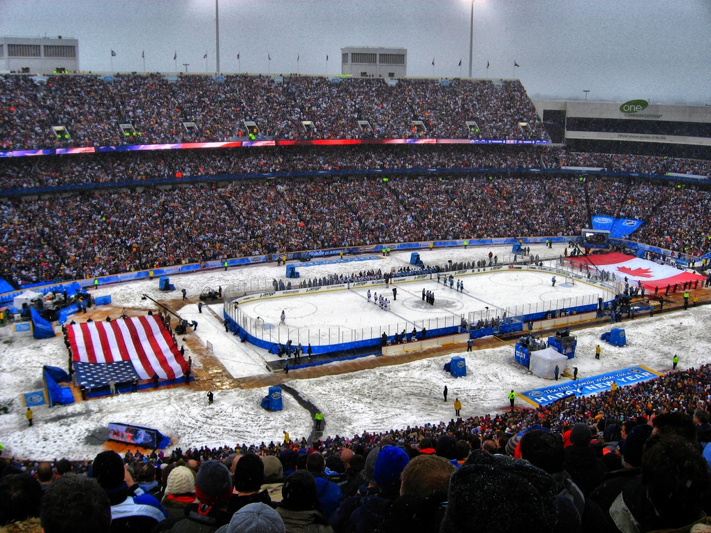Outdoor NHL Winter Classic game being played in a snow-filled stadium crowded with fans, featuring a large American flag during the opening ceremony.