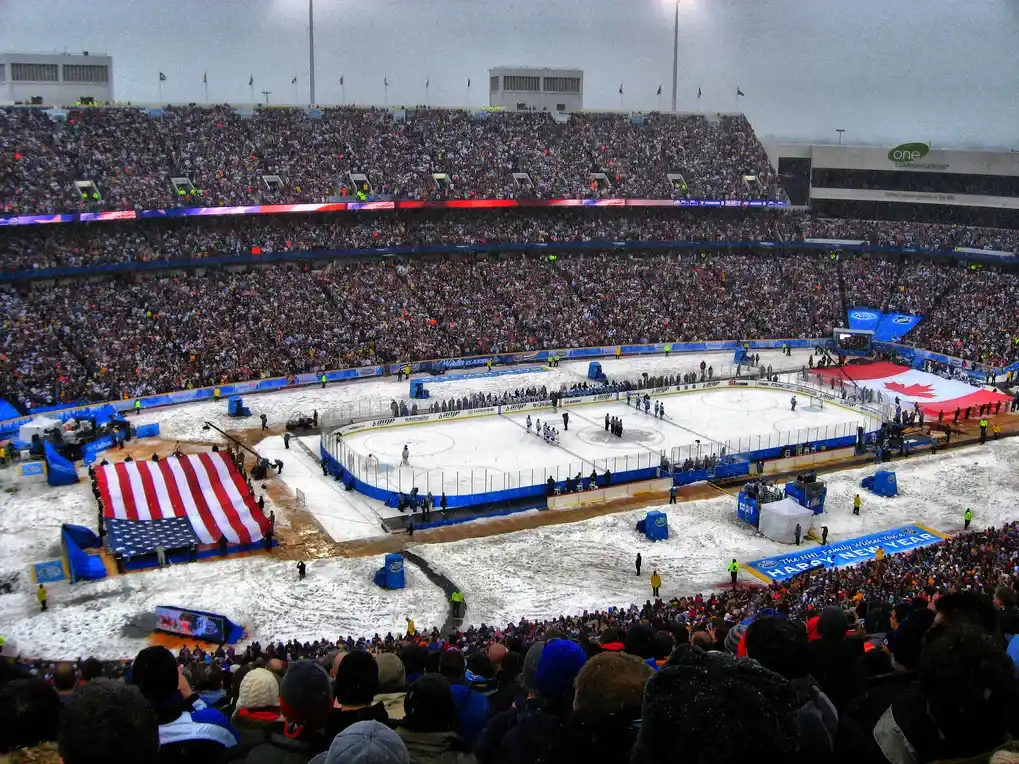 Outdoor NHL Winter Classic game being played in a snow-filled stadium crowded with fans, featuring a large American flag during the opening ceremony.