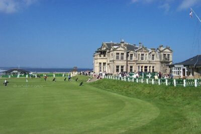 Golfers play on one of the top British Open golf courses, which features a historic clubhouse and well-maintained greens under a clear sky.