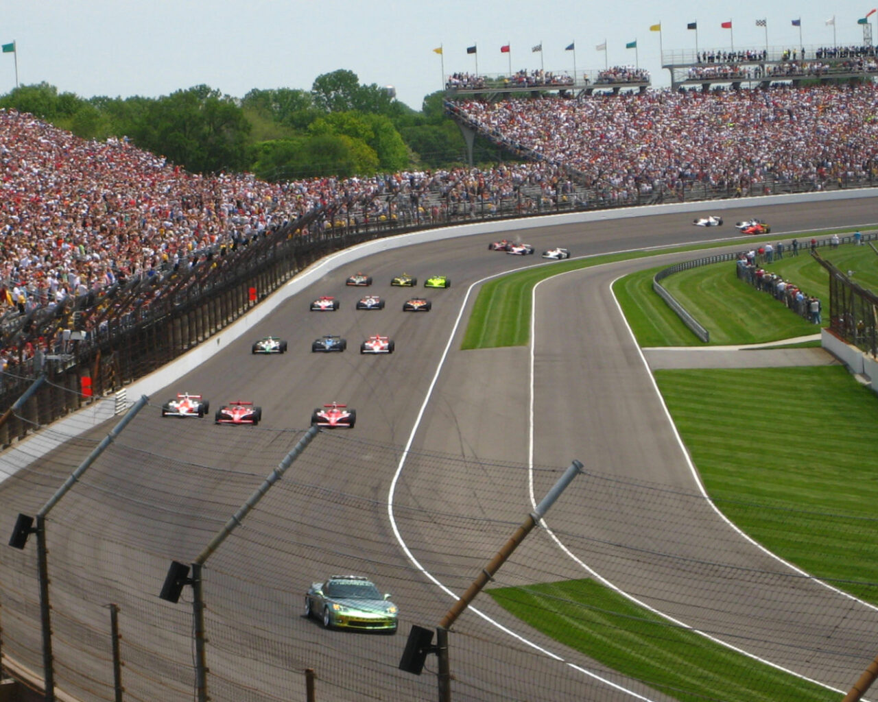 Race cars speed along the track at the Indianapolis Motor Speedway during the Indy 500, with a large crowd watching from the packed grandstands.