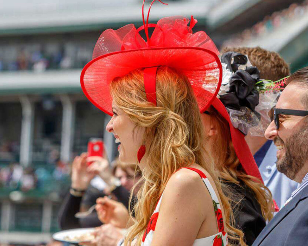 Woman at the Kentucky Derby wearing a striking red hat, enjoying the event with others visible in the background, reinforcing the festive atmosphere of the Kentucky Derby Packages.