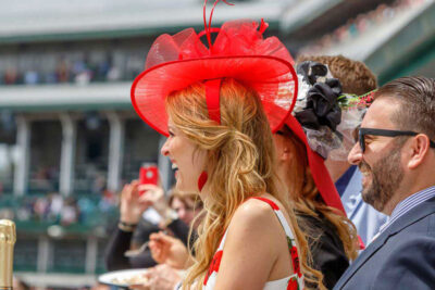 Woman at the Kentucky Derby wearing a striking red hat, enjoying the event with others visible in the background, reinforcing the festive atmosphere of the Kentucky Derby Packages.