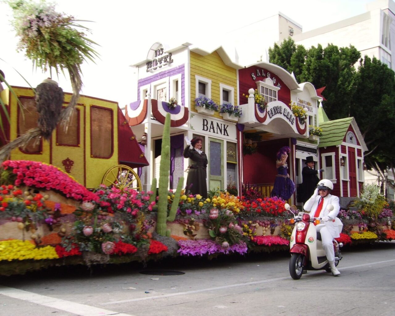A float adorned with vibrant flowers displaying a vintage town theme passes by during a parade, accompanied by a person riding a white scooter, enhancing the festive atmosphere of the Rose Bowl event.