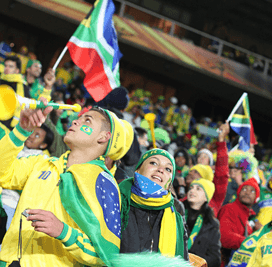 Two soccer fans, wearing Brazil colors and holding a Brazilian flag, cheer excitedly at a sports event, embodying the vibrant atmosphere highlighted in Sports Travel Blog by Roadtrips.
