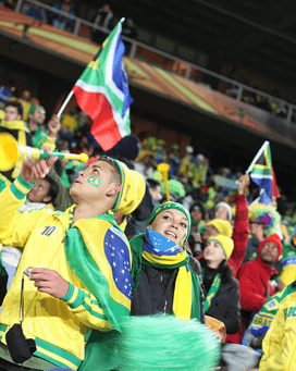 Two soccer fans, wearing Brazil colors and holding a Brazilian flag, cheer excitedly at a sports event, embodying the vibrant atmosphere highlighted in Sports Travel Blog by Roadtrips.