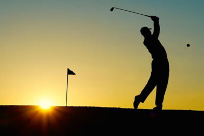 Silhouetted golfer swings at a golf ball near a flag at sunset, illustrating a classic golf trip moment.