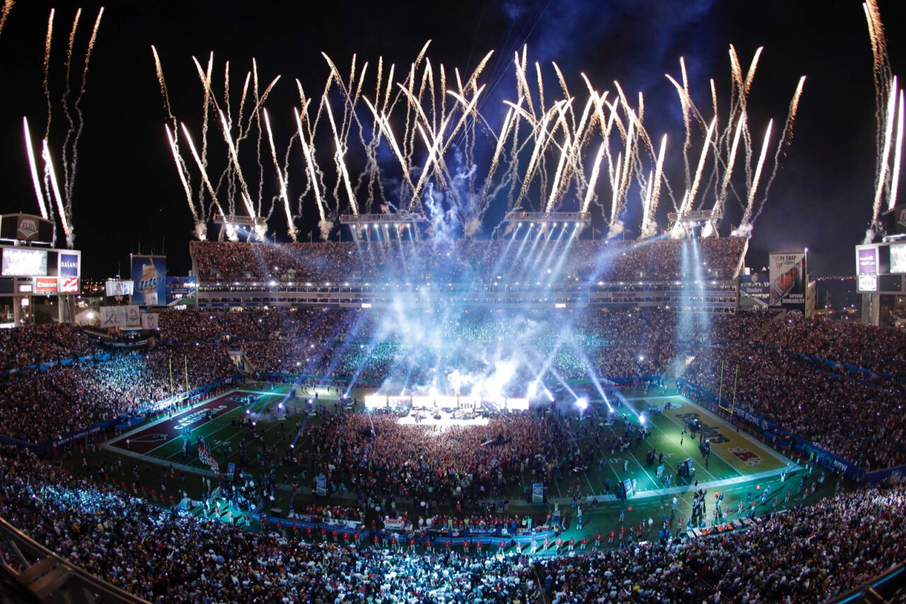 Fireworks illuminate the stadium during a Super Bowl event, filled with a large crowd and vibrant light effects.