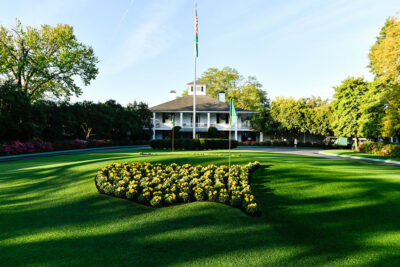 Sunlight bathes the iconic clubhouse at Augusta National, framed by immaculate gardens and a floral display shaped like the United States, capturing a classic scene at The Masters.