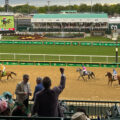 Spectators cheer on jockeys and horses racing at the Kentucky Derby, set against the backdrop of the track's iconic grandstands and a large display screen showing the race.