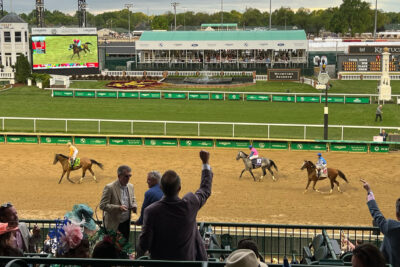Spectators cheer on jockeys and horses racing at the Kentucky Derby, set against the backdrop of the track's iconic grandstands and a large display screen showing the race.