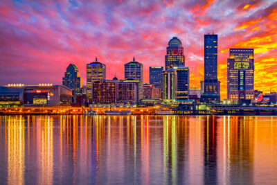 Vibrant sunset colors reflect off the Ohio River behind the Louisville skyline, highlighting sites like the KFC Yum! Center, making it a picturesque setting during events like the Kentucky Derby.