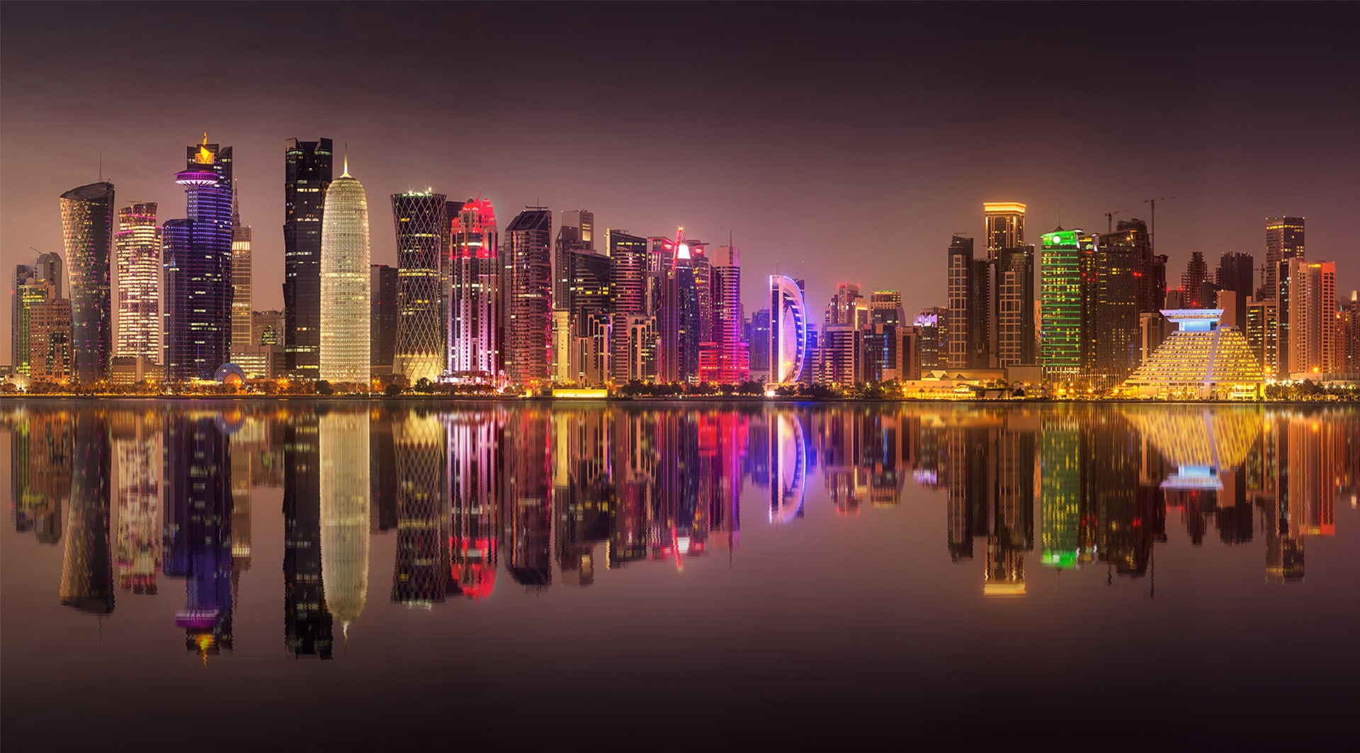 A vibrant nighttime view of Doha's skyline reflecting in the water, showcasing a range of illuminated modern buildings and towers.