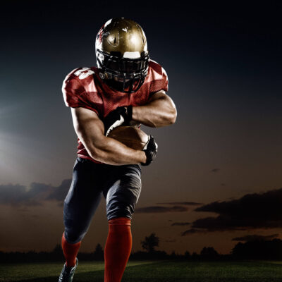 Football player in red uniform clutching a ball, showcasing a Super Bowl handoff play against a dusk sky backdrop.