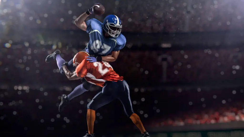 A football player in a blue uniform jumps and clutches the football while dodging a tackle during a nighttime game.