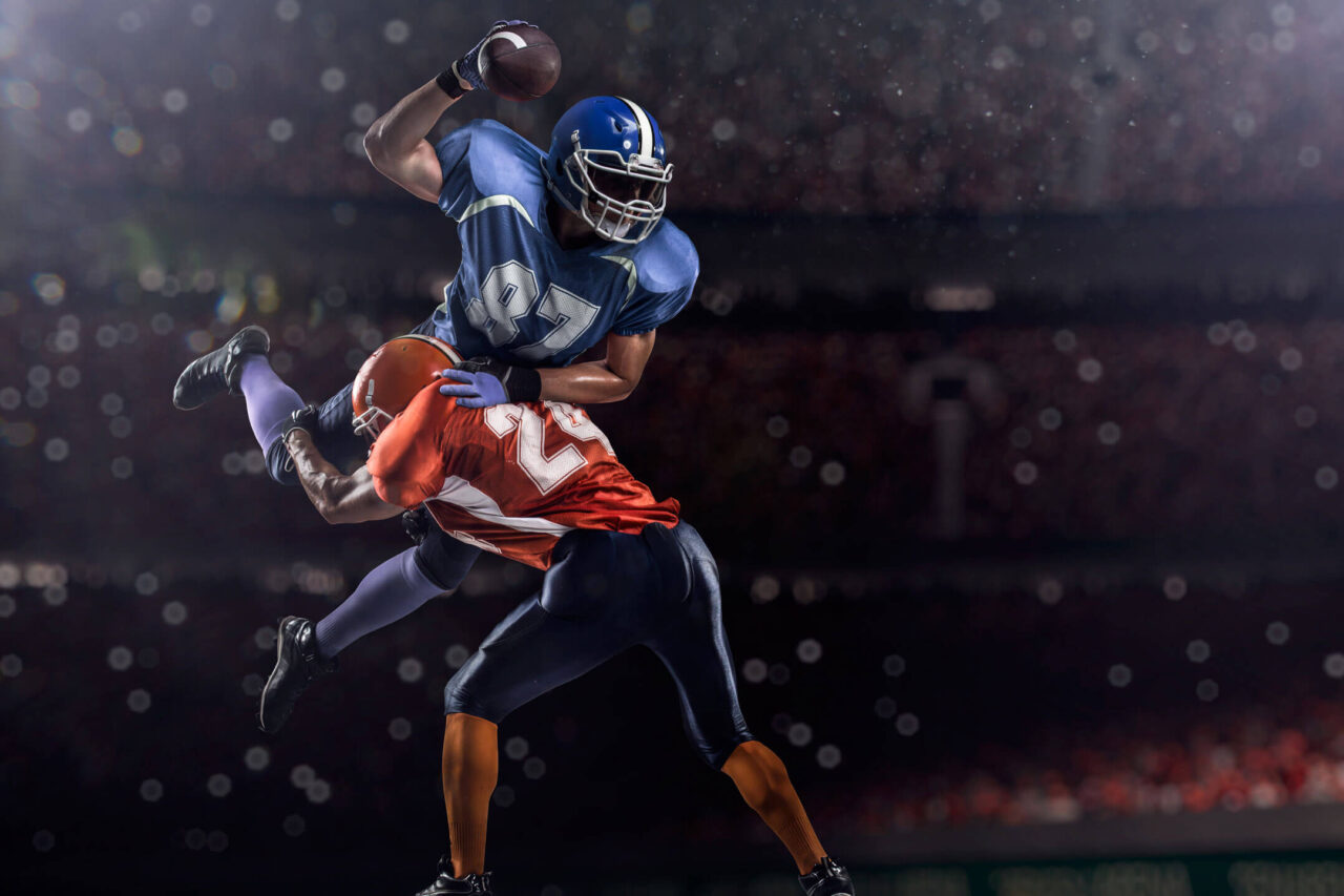 Football player in blue jumps with the ball, avoiding a tackle from an opponent in orange during a vibrant night game, capturing the thrill of football travel packages.