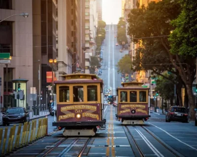 Two cable cars travel on tracks up a steep street in San Francisco, surrounded by buildings and trees.