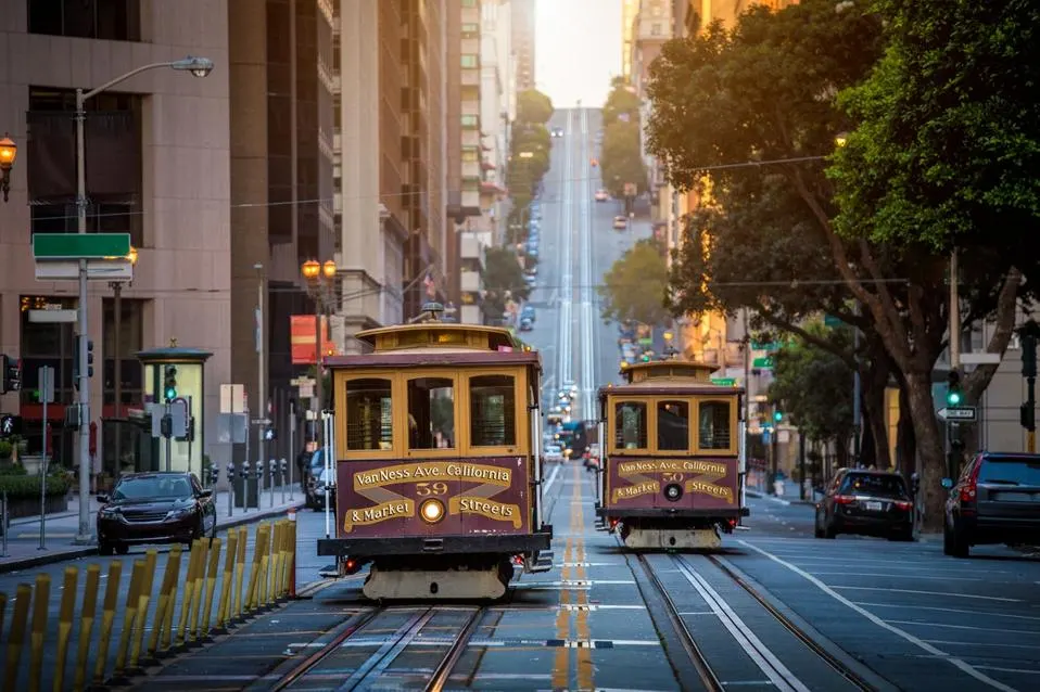 Two cable cars travel on tracks up a steep street in San Francisco, surrounded by buildings and trees.