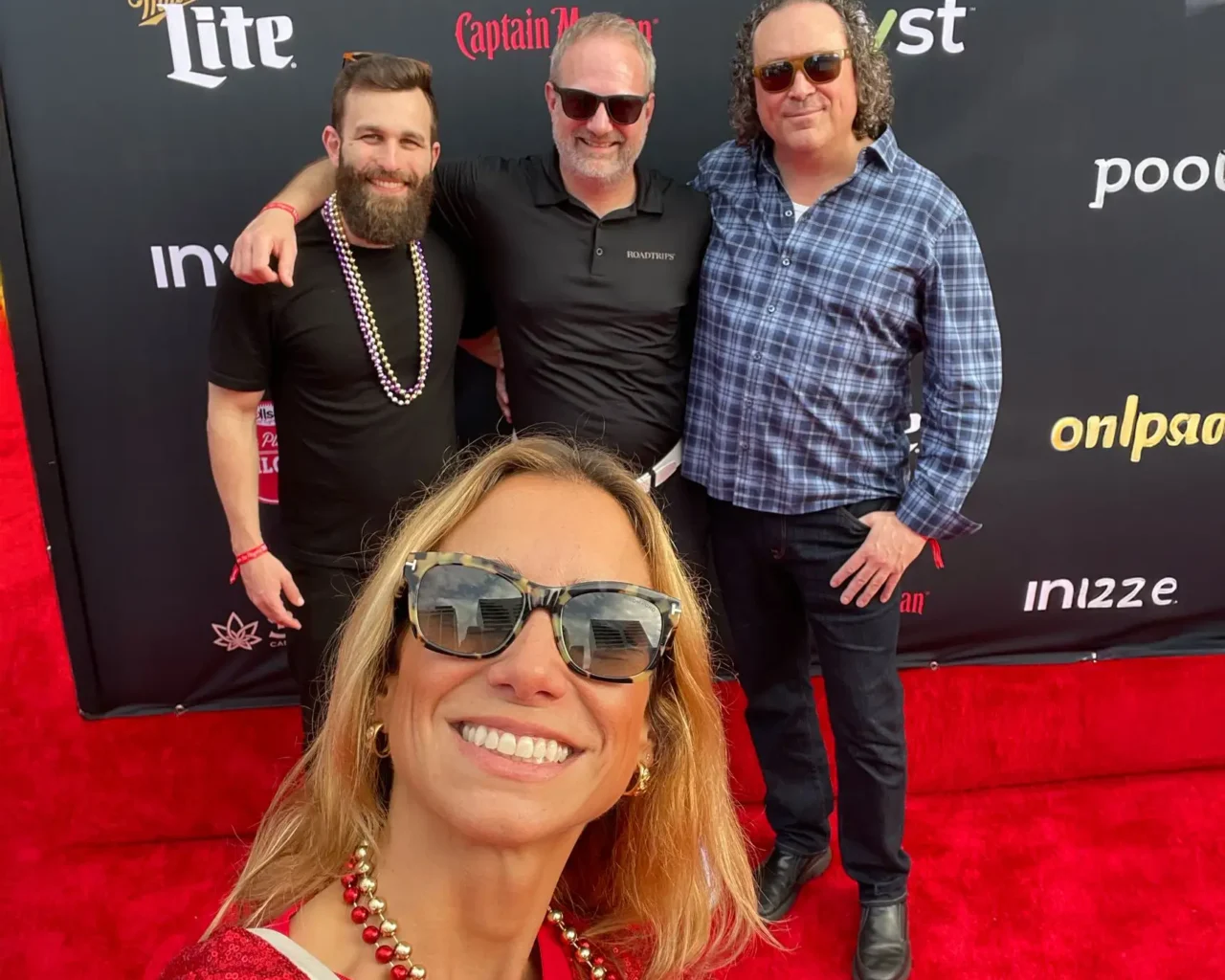Four people pose together on a red carpet event, featuring a woman in the foreground taking a selfie with three men smiling in the background.