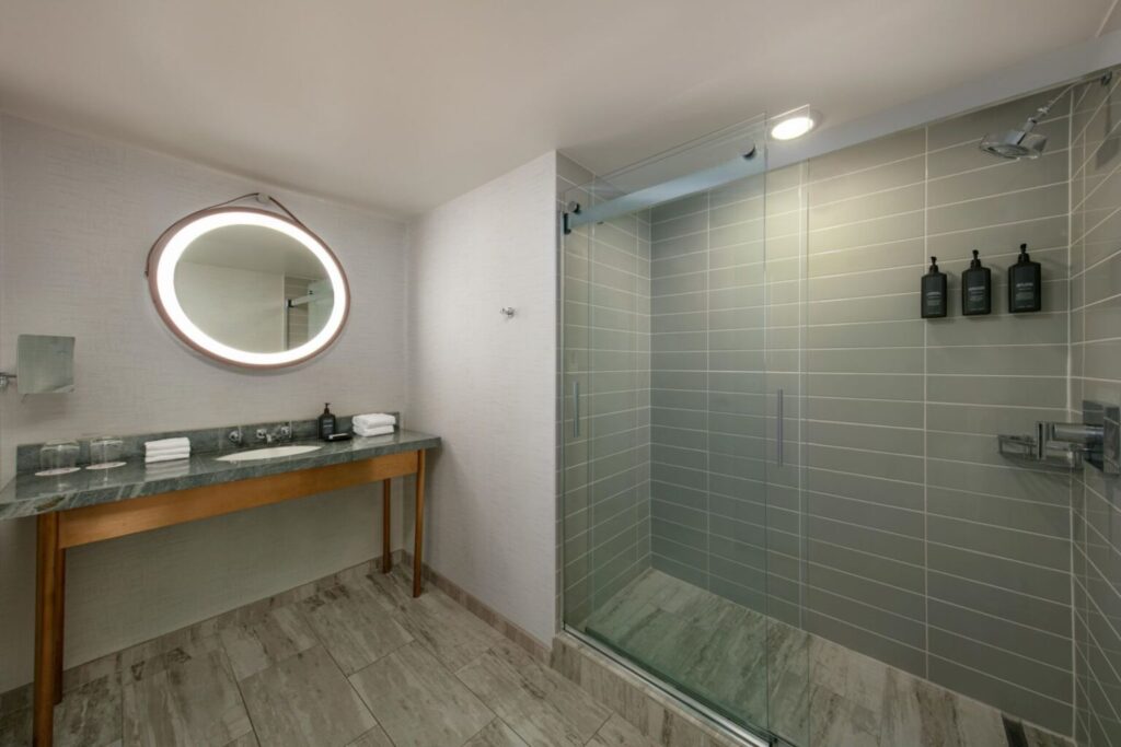 Minimalist bathroom interior featuring a wall-mounted mirror with LED lighting, wooden vanity, and a spacious walk-in shower, located at Hotel Dena near the Rose Bowl in Pasadena, California.
