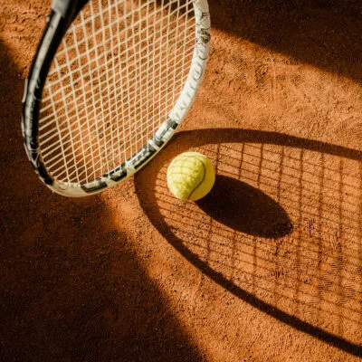 Close-up of a tennis ball and racquet on a clay court, highlighting the texture of the surface and shadows cast by the sunlight, ideal for luxury sports travel enthusiasts.