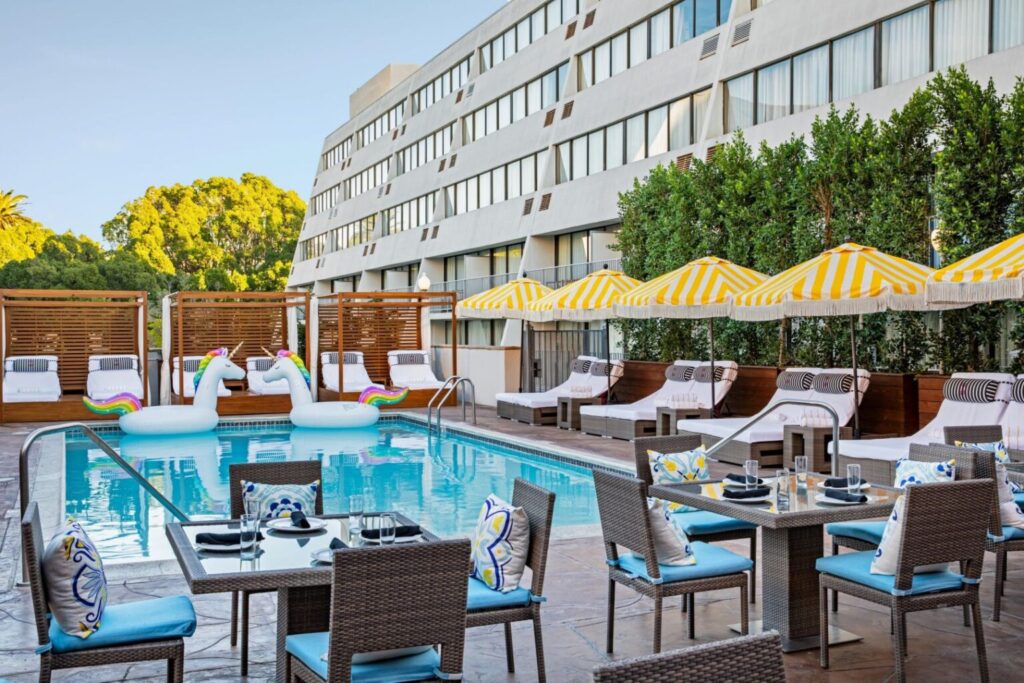 Luxury outdoor pool area at Hotel Dena in Pasadena, featuring striped yellow umbrellas, lounge chairs, and playful unicorn pool floats, ideal for Rose Bowl visitors.