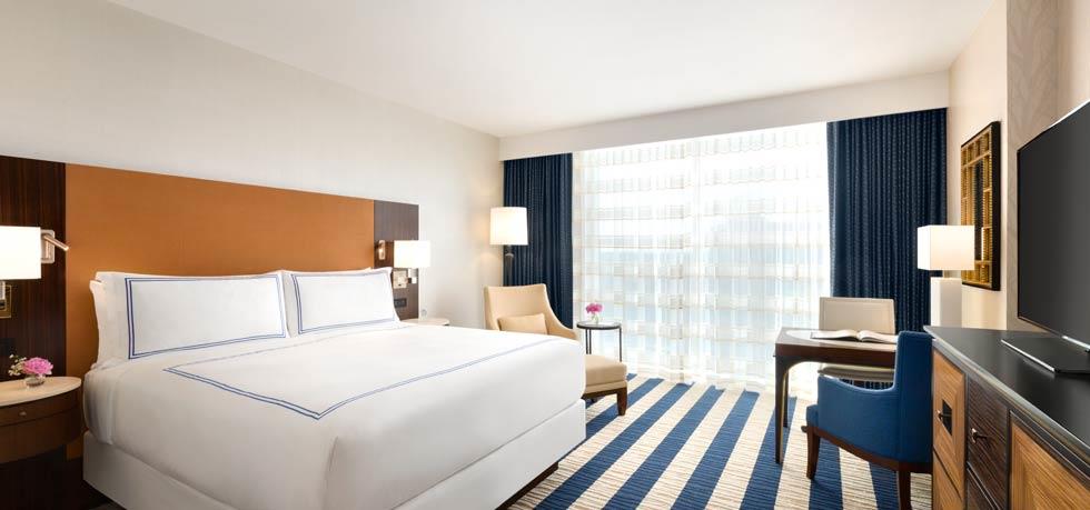 A luxurious hotel room featuring a neatly made bed with a brown headboard, a seating area, and expansive windows, suggestive of accommodations for the US Grand Prix in Austin, Texas.
