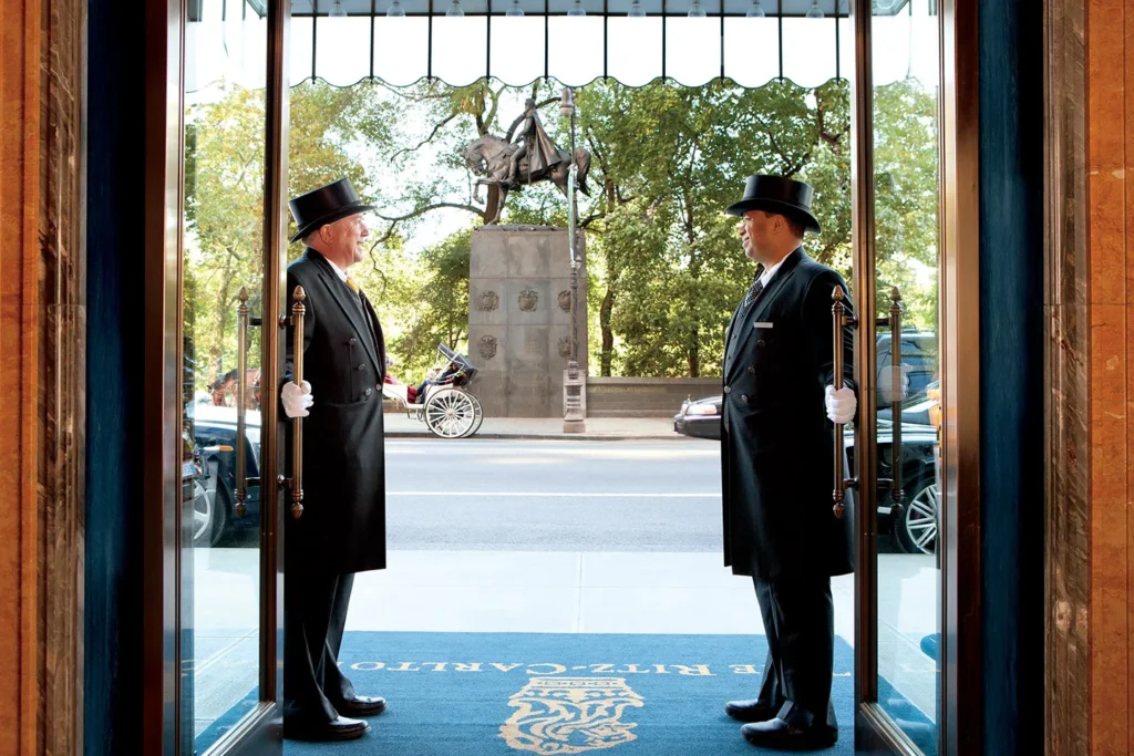 Two doormen in traditional uniforms stand at the open entrance of the Ritz-Carlton Central Park, overlooking a busy New York street with a view of Central Park.