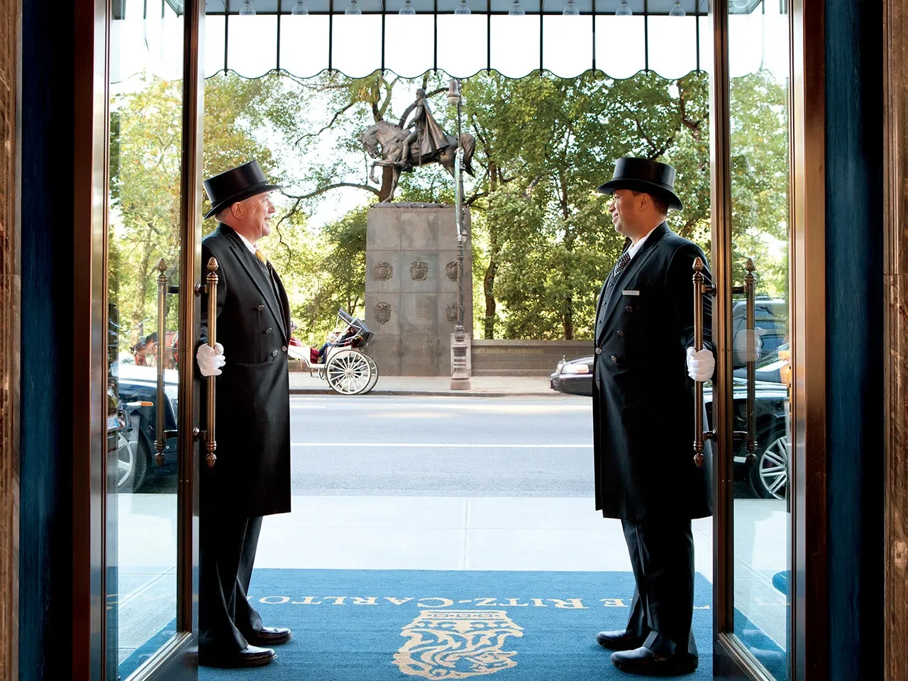 Two doormen in traditional uniforms stand at the open entrance of the Ritz-Carlton Central Park, overlooking a busy New York street with a view of Central Park.