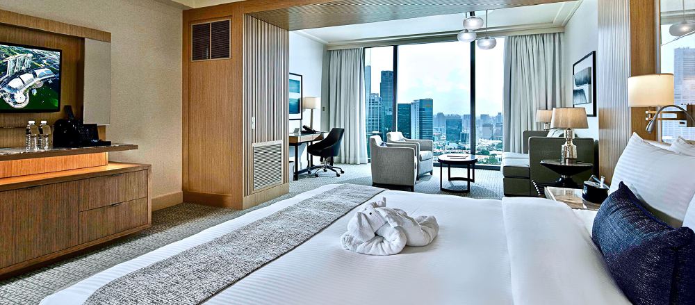 Luxury hotel room overlooking the cityscape of Singapore, featuring a large bed, a seating area, and a wall-mounted TV showing a sports event, ideal for guests attending the Singapore Grand Prix.