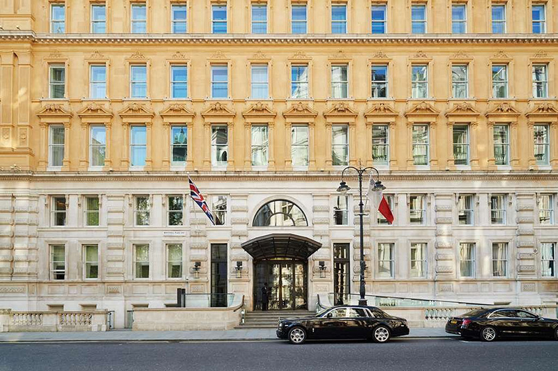 Exterior view of the Corinthia Hotel in London, showing its grand entrance flanked by flags and parked cars in front.