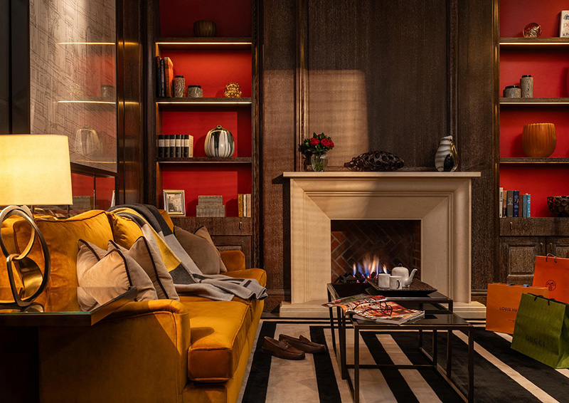 Luxury lounge area in Corinthia Hotel London featuring an inviting fireplace, plush yellow sofa, contemporary decor, and upscale shopping bags, ideal for guests attending Wimbledon.