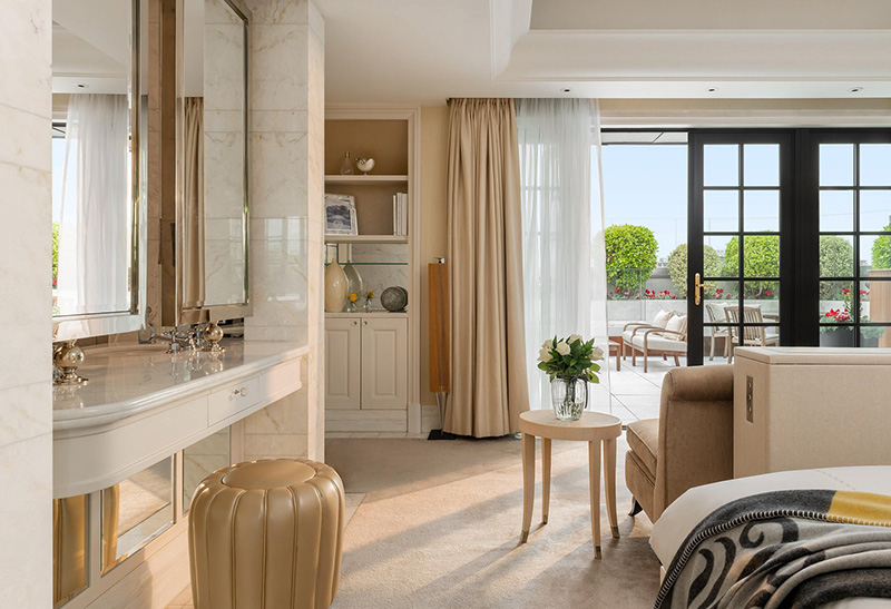 Luxurious suite in the Corinthia Hotel London, featuring a marble vanity, plush seating, and doors leading to a sunlit balcony, ideal for guests attending Wimbledon Championships.