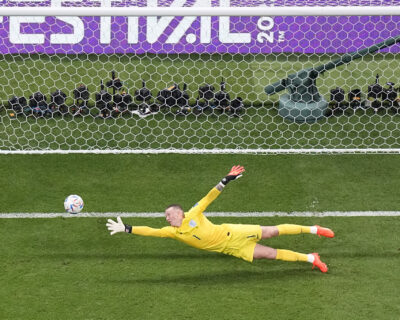 Soccer goalkeeper in yellow dives towards the ball during a match, with a view of the goal and photographers in the background, relevant for discussions about luxury travel packages to major sports events.