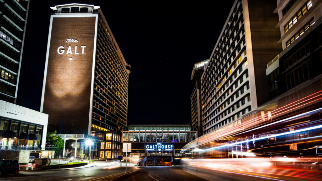 Nighttime view of The Galt House Hotel in Louisville, Kentucky, illuminated with light trails from moving vehicles in the foreground.
