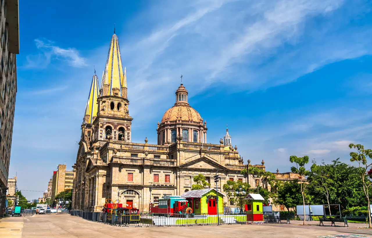 Colorful buildings and a playground sit in front of Guadalajara's iconic cathedral under a clear blue sky, highlighting its status as a World Cup host city with Estadio Akron.