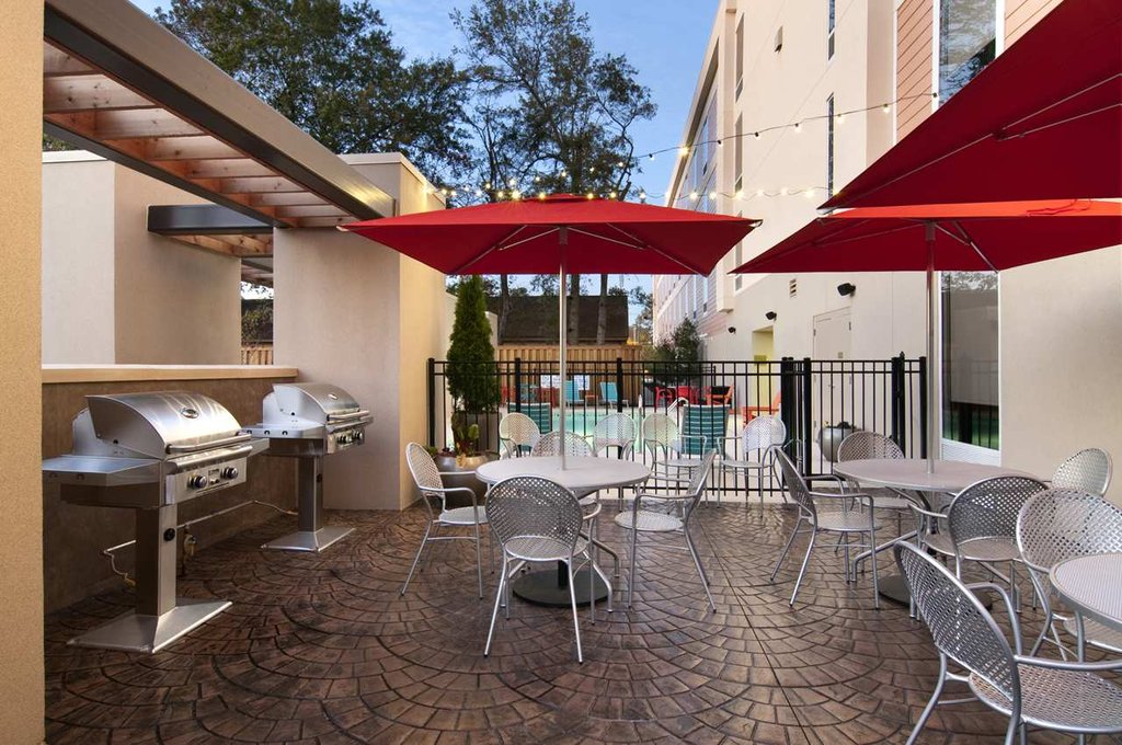 Outdoor grilling patio at Hilton Home2 Suites in Augusta, Georgia, featuring two large grills under a pergola, multiple seating areas with red umbrellas, and a view of the pool area.