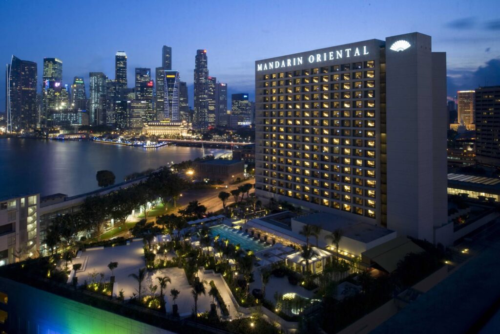 Twilight view of the Mandarin Oriental Hotel in Singapore, prominently situated foreground with a vibrant cityscape and bay in the background, ideal for visitors attending the Singapore Grand Prix.