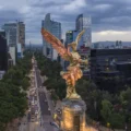 Overlooking Mexico City, the golden Angel of Independence statue stands prominently above a bustling avenue flanked by modern skyscrapers, highlighting the city's status as a host for the World Cup.