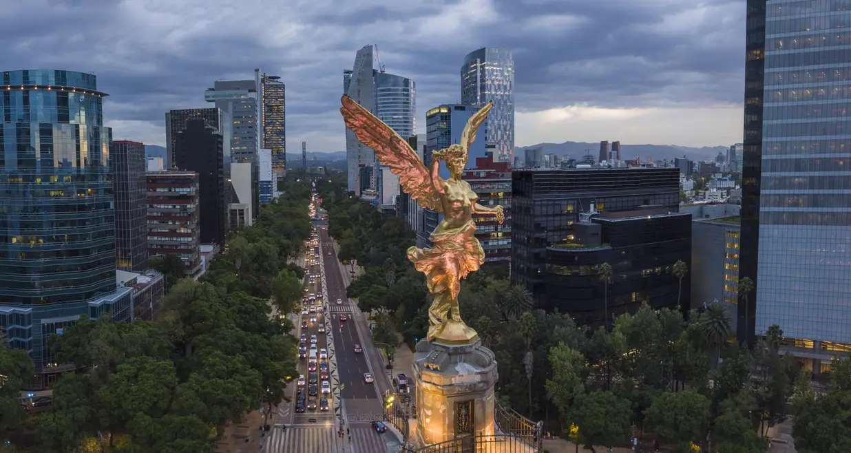 Overlooking Mexico City, the golden Angel of Independence statue stands prominently above a bustling avenue flanked by modern skyscrapers, highlighting the city's status as a host for the World Cup.
