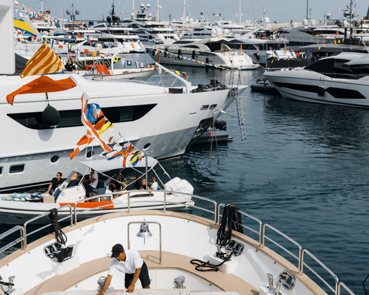 Luxury yachts decorated with colorful flags are moored in a bustling marina, likely part of the Monaco Grand Prix viewing and hospitality packages, with people and crew visible on deck.