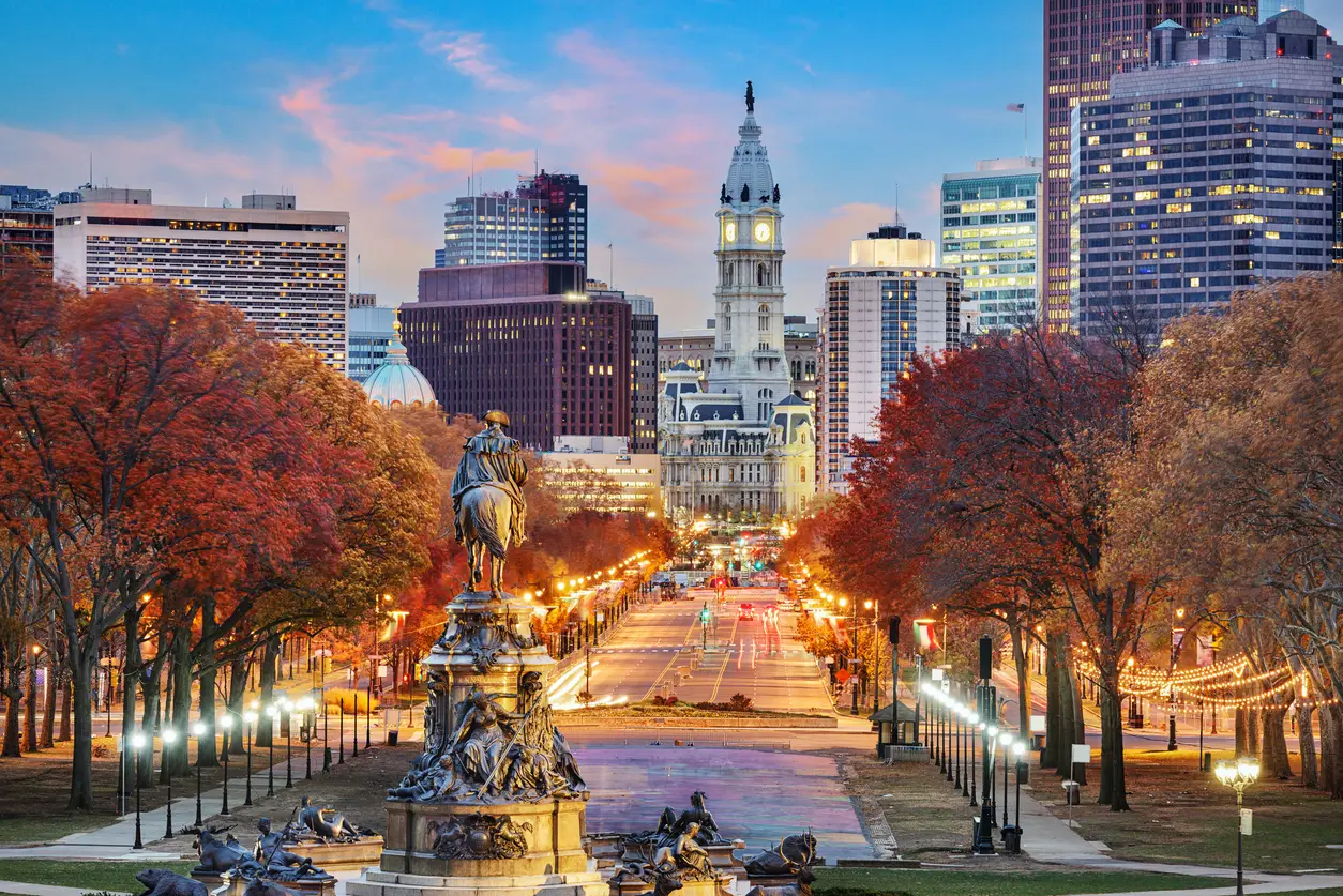 View of Philadelphia featuring Benjamin Franklin Parkway leading up to the historic City Hall, illuminated during twilight, with autumn-colored trees and a statue in the foreground.