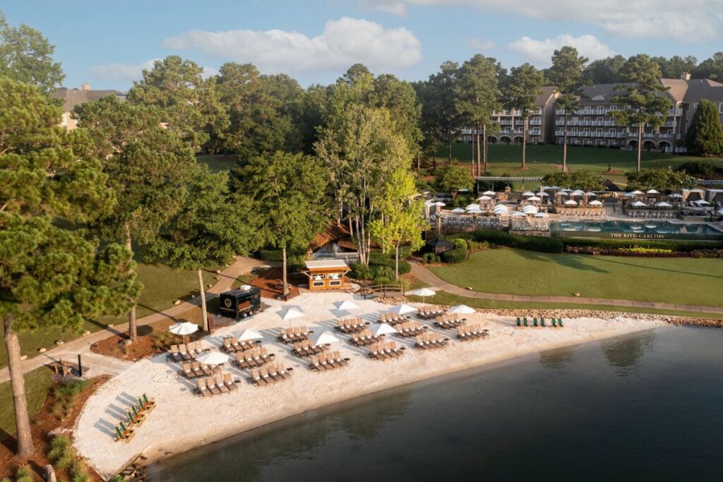 Aerial view of the Ritz-Carlton Reynolds at Lake Oconee, showing beach chairs lined up along the sandy shore, adjacent to a lush green lawn and hotel buildings. Ideal for travelers seeking accommodations near Augusta, GA.