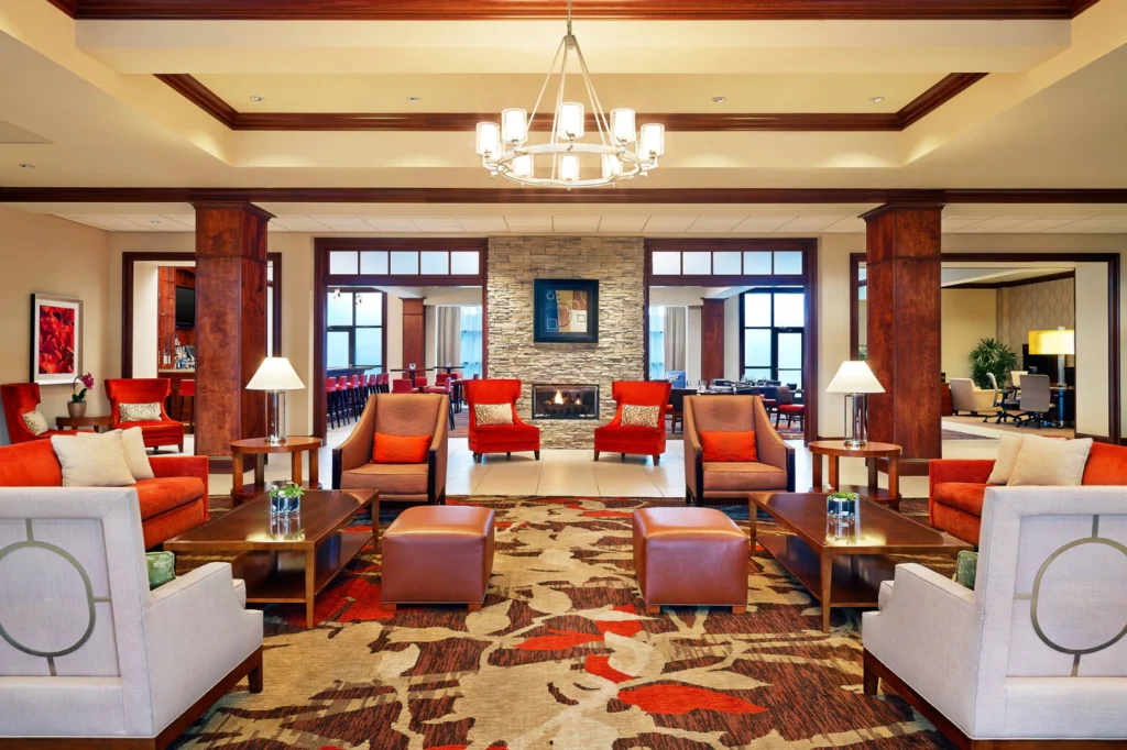 Warmly decorated lobby at the Sheraton Augusta Hotel, featuring red and orange hues, plush seating, wood accents, and a welcoming fireplace, ideal for Masters Hotels in Augusta, GA.