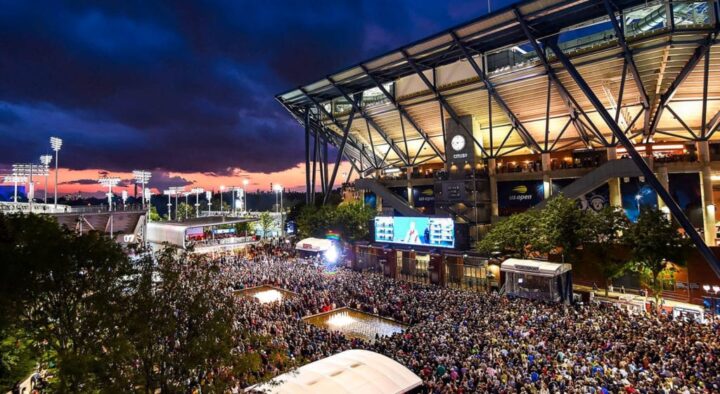 A bustling evening crowd gathers at the Arthur Ashe Stadium for the US Open Tennis Schedule event under a dramatic sunset sky.