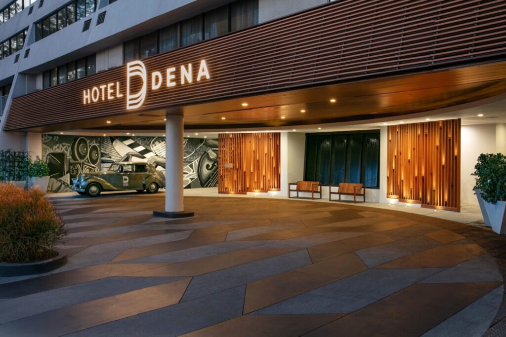 Elegant entrance of Hotel Dena in Pasadena, showcasing its illuminated signage, stylish bench seating, and a vintage car on display under a wooden paneled overhang. Ideal for Rose Bowl accommodations.