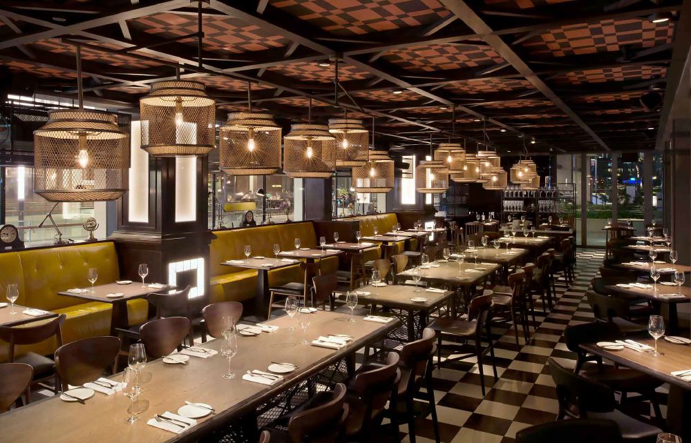 Interior of Bread Street Kitchen in Singapore, showcasing elegantly set tables, stylish oversized wicker lamps, and a distinctive black and white tiled floor, located near Marina Bay Sands. Ideal for visitors to luxury hotels in Singapore, such as those attending the Singapore Grand Prix.
