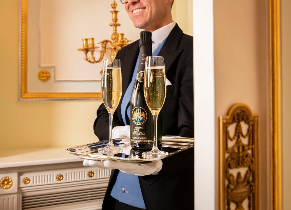 A butler in a smart suit serves champagne at the Ritz London, reflecting the luxury accommodations available for visitors during the Wimbledon Championships.